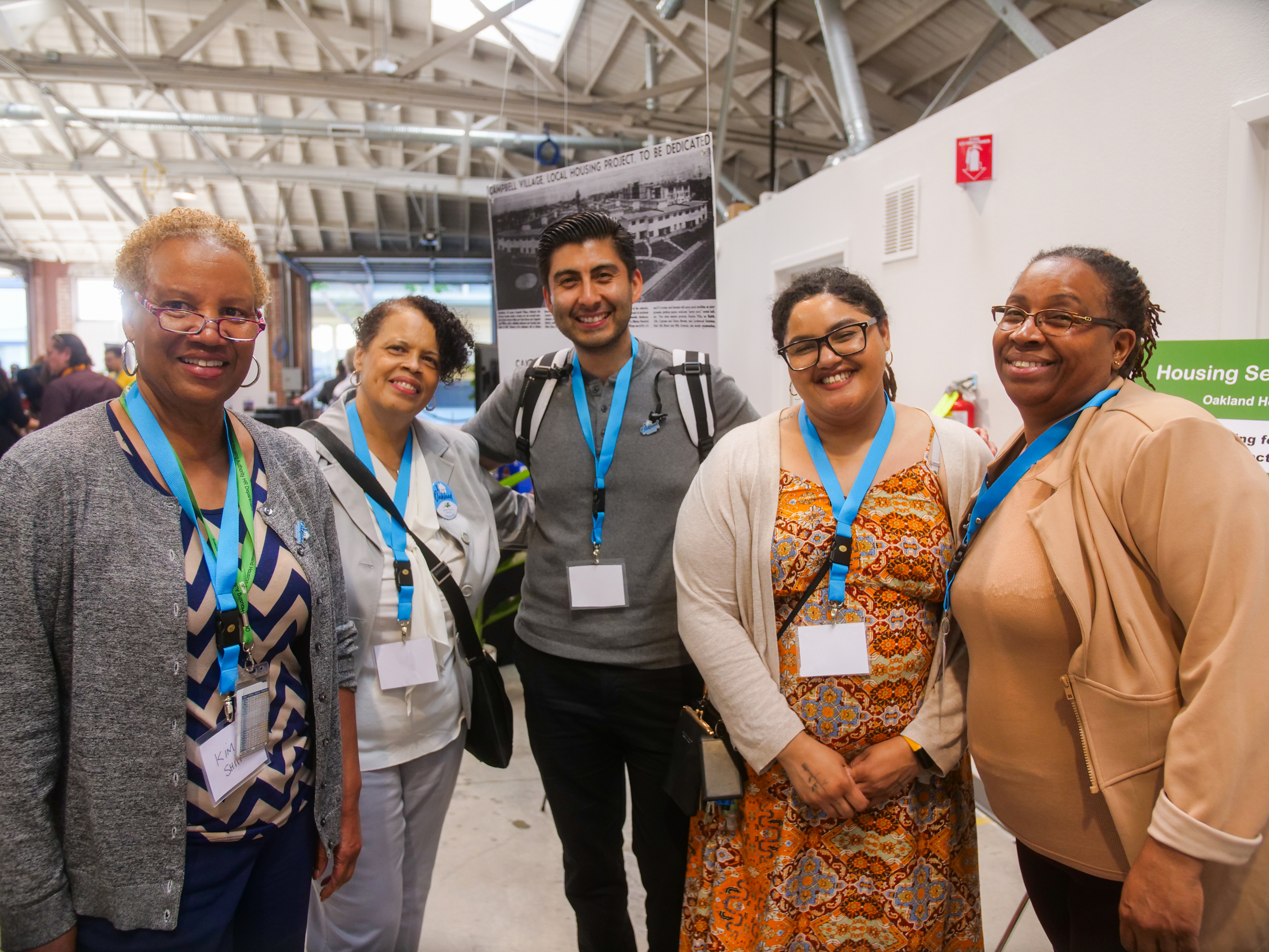 A group of five people smiling, wearing lanyards and name tags at an indoor event. Bright setting with a modern industrial background.