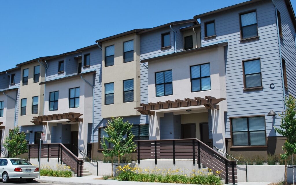 Modern three-story townhouses with beige and gray exteriors under a clear blue sky. Manicured landscaping and a parked car convey a suburban setting.