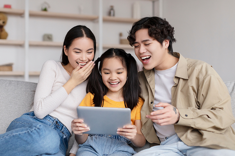 Laughing young asian husband, wife and teen daughter watching funny video on tablet, surfing in internet, sits on sofa in living room interior, copy space. Modern device, app, website and online game