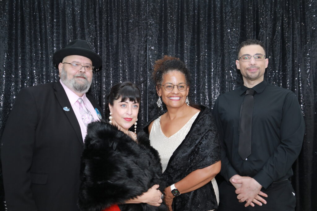 Four Oakland Housing Authority staff members pose for a photo at Senior Splendor 2025 in front of a black sequin backdrop. Executive Director Patricia Wells is second from right.