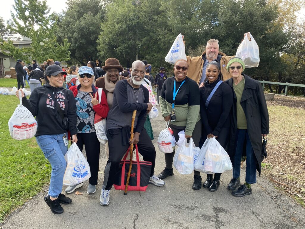 A group of smiling volunteers stand together holding bags of food at an outdoor holiday distribution event.