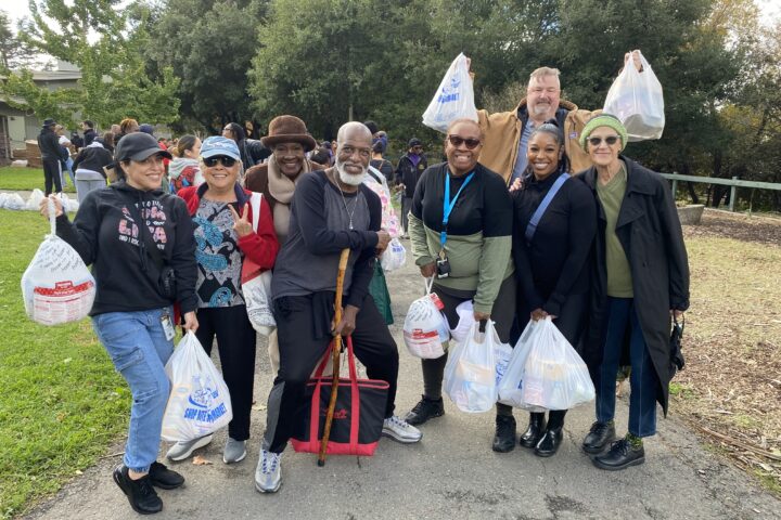 Alt Text: A group of OHA staff and residents smiling while holding holiday meal bags at the District 6 Thanksgiving Food Giveaway.