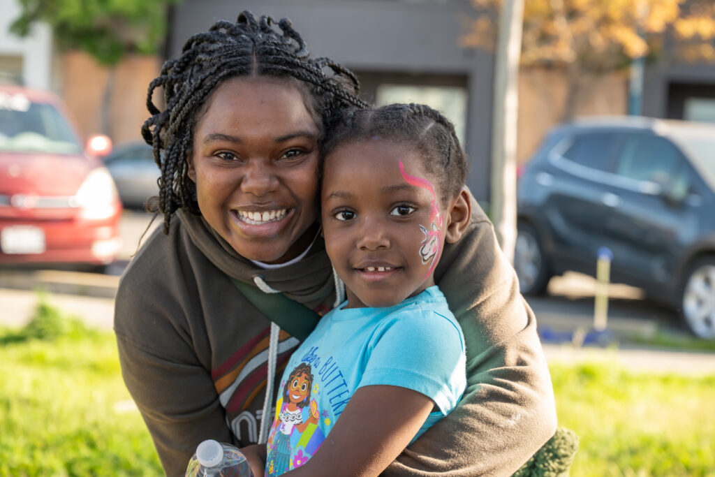 A smiling adult embraces a young girl with face paint at an outdoor community gathering.