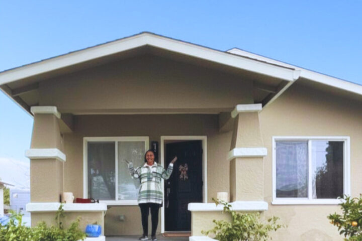 A resident stands on the porch of her new East Oakland home with arms raised in celebration.