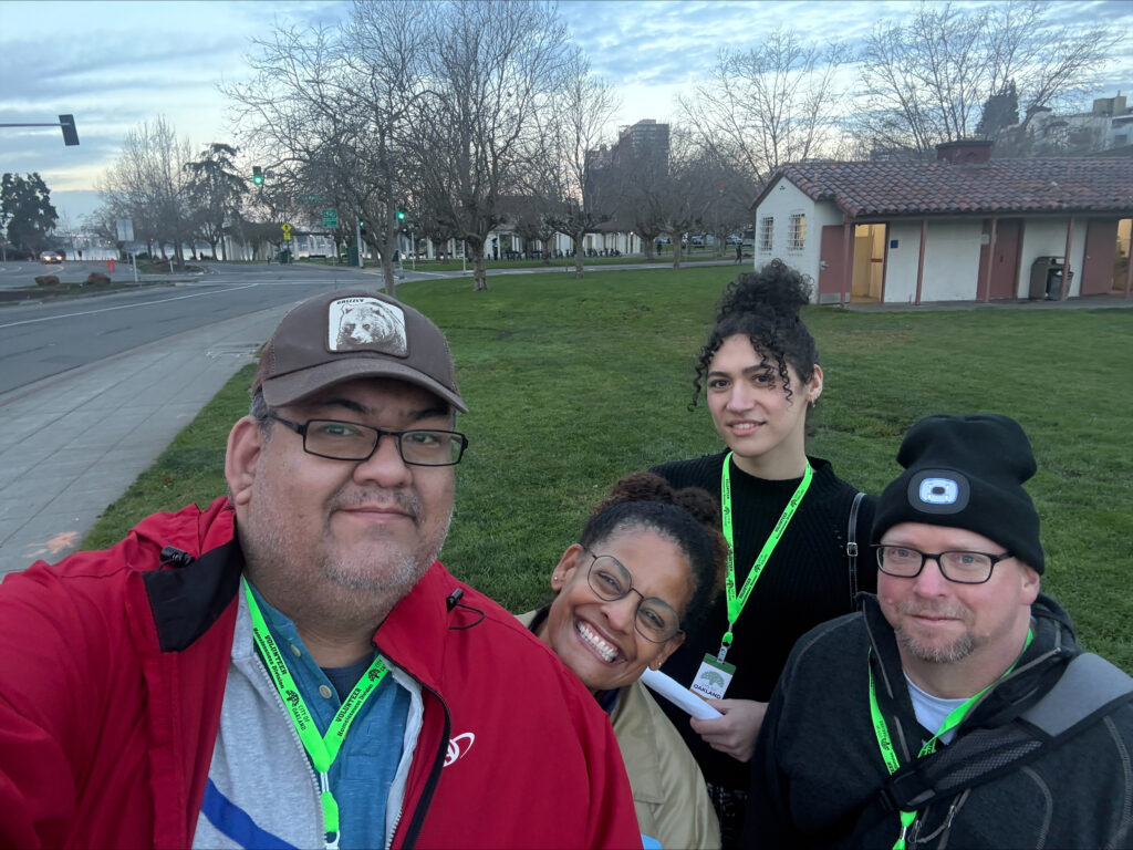 Four Oakland Housing Authority (OHA) Point-in-Time (PIT) count volunteers wearing green lanyards smile for a selfie in an Oakland park on an overcast day.