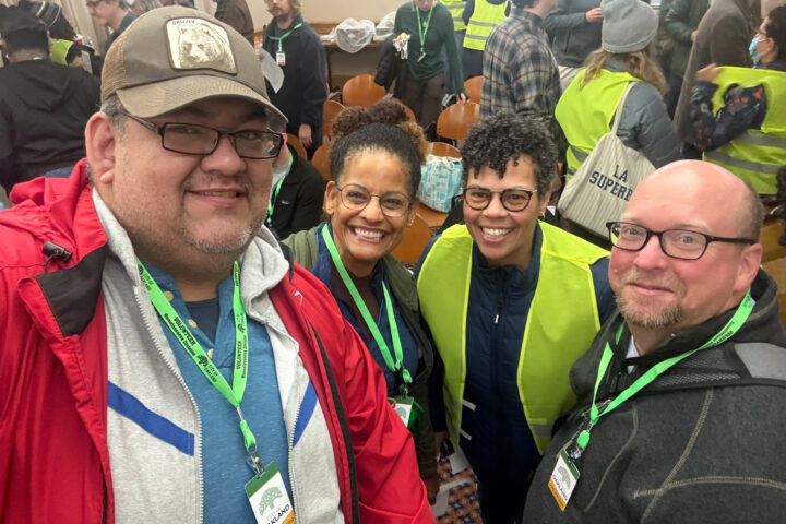 Four Oakland Housing Authority (OHA) Point-in-Time (PIT) count volunteers wearing green lanyards smile for a selfie in a busy room full of volunteers.