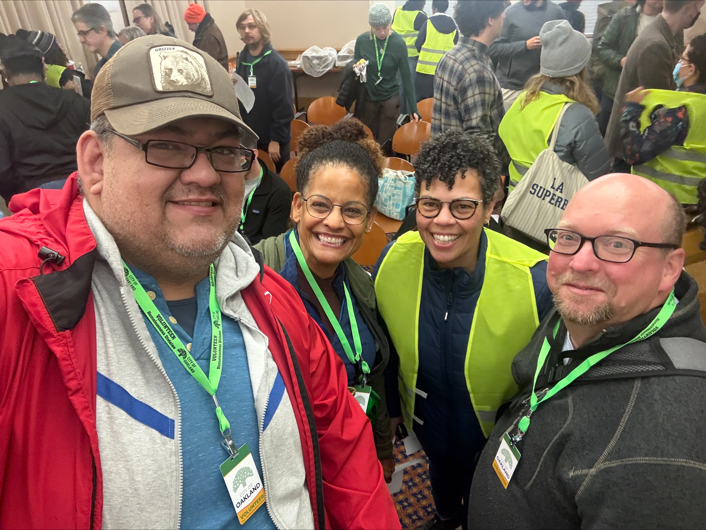 Four Oakland Housing Authority (OHA) Point-in-Time (PIT) count volunteers wearing green lanyards smile for a selfie in a busy room full of volunteers.
