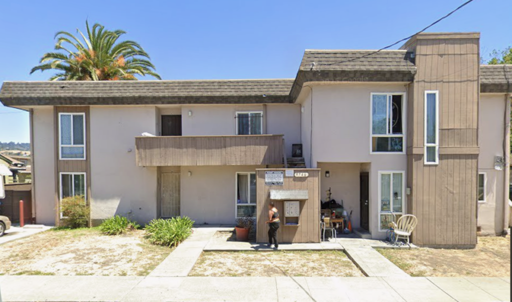 Exterior view of a multi-unit residential building in East Oakland before rehabilitation, showing worn siding and aging structural elements.