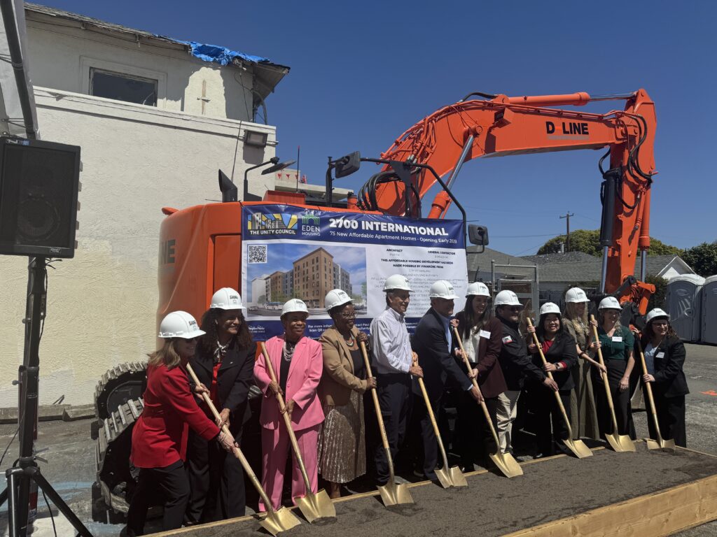 Community partners stand with ceremonial shovels at the groundbreaking for 2700 International Blvd. in Oakland, with construction equipment and a project sign in the background.